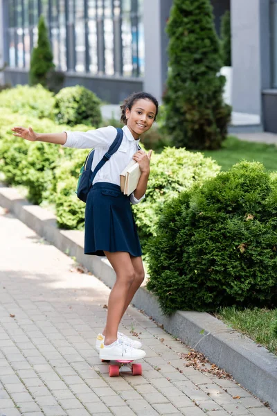 Feliz Africano americano estudante com mochila e livros montando penny board ao ar livre — Fotografia de Stock