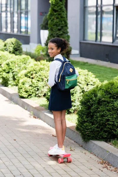 Souriant afro-américaine écolière avec sac à dos équitation penny board à l'extérieur — Photo de stock