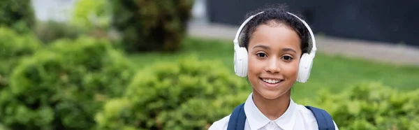 Sonriente afroamericana colegiala en auriculares al aire libre, tiro panorámico - foto de stock