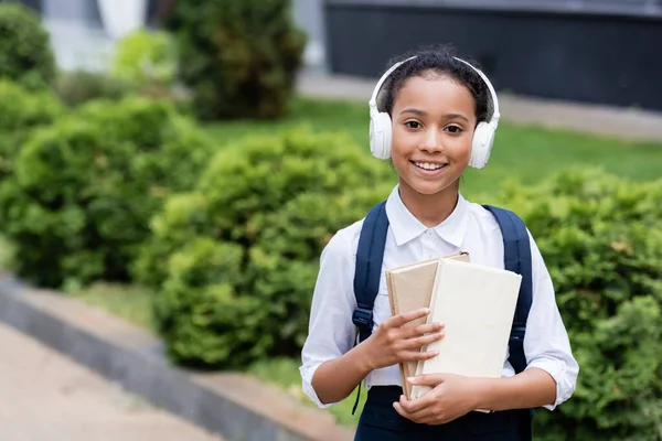 Smiling african american schoolgirl in headphones with backpack and books outdoors — Stock Photo