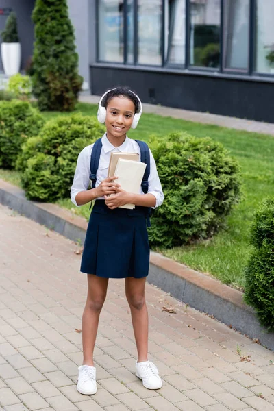 Smiling african american schoolgirl in headphones with backpack and books outdoors — Stock Photo