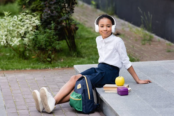 Souriant afro-américaine écolière écouter de la musique dans les écouteurs près du sac à dos, livres et déjeuner en plein air — Photo de stock