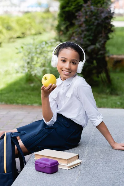 Colegiala afroamericana sonriente en auriculares con manzana cerca de la mochila, libros y lonchera al aire libre - foto de stock