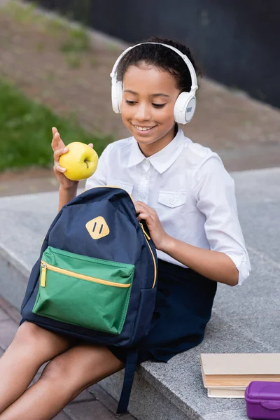 Smiling african american schoolgirl in headphones holding apple and backpack near books and lunch box outdoors — Stock Photo