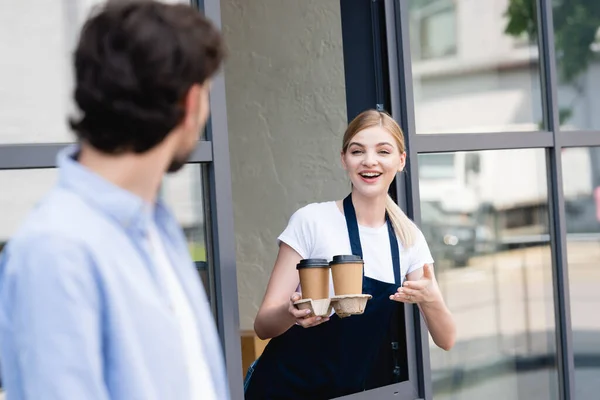 Foco seletivo de garçonete sorridente apontando com a mão para o café para ir ao homem na rua urbana — Fotografia de Stock