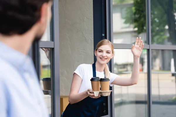 Enfoque selectivo de camarera sonriente sosteniendo vasos de papel y saludando al hombre cerca de la cafetería - foto de stock