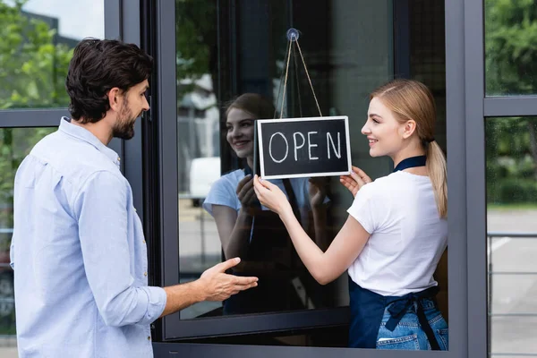 Customer pointing with hand near smiling waitress holding signboard with open lettering on door of cafe — Stock Photo