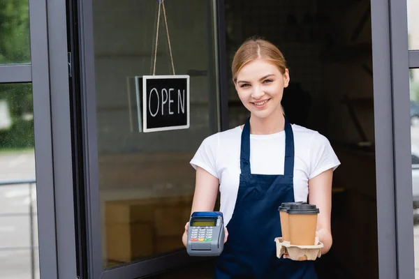 Усміхнена офіціантка тримає платіжний термінал і каву, щоб підійти до вивіски з відкритою пляшкою на дверях кафе — стокове фото