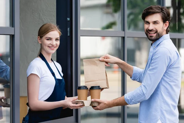 Garçonete sorridente e cliente segurando café para ir e saco de papel enquanto sorri para a câmera perto de café na rua urbana — Fotografia de Stock