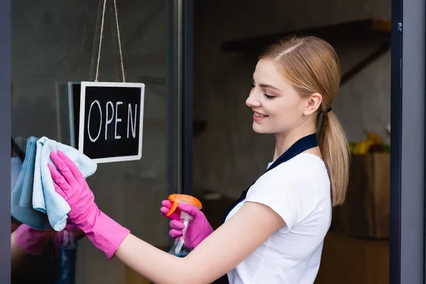 Vue latérale de la serveuse souriante nettoyage porte du café près de l'enseigne avec lettrage ouvert — Photo de stock