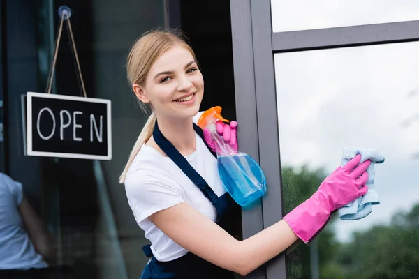 Cameriera sorridente in uniforme finestra di pulizia del caffè vicino al cartello con scritte aperte sulla porta — Foto stock