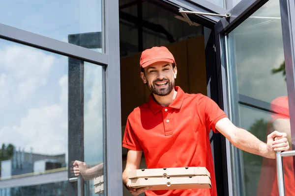 Camarero sonriente sosteniendo cajas de pizza cerca de café en la calle urbana - foto de stock