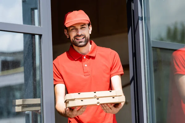 Serveur souriant en uniforme tenant des boîtes à pizza près d'un café dans une rue urbaine — Photo de stock