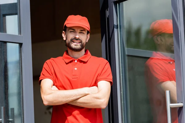 Handsome waiter in uniform with crossed arms smiling at camera near cafe on urban street — Stock Photo