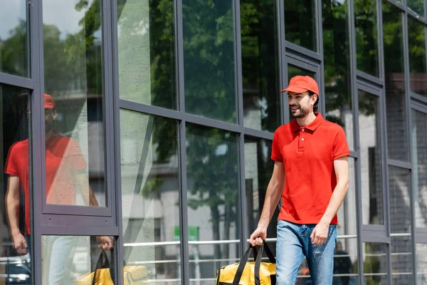 Homem de entrega sorridente segurando saco térmico enquanto caminhava perto do edifício na rua urbana — Fotografia de Stock
