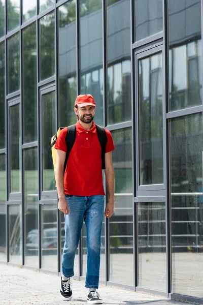 Smiling delivery man in uniform and thermo backpack walking on urban street — Stock Photo