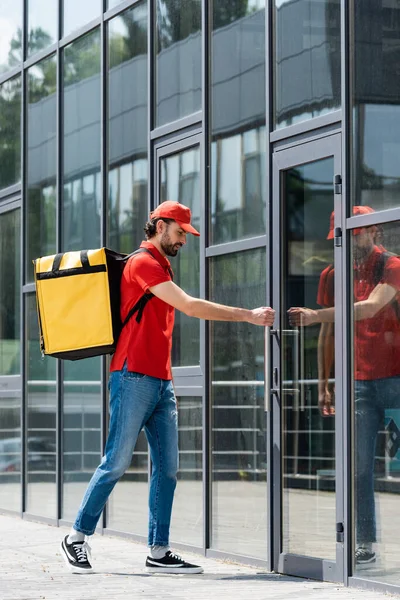Delivery man in uniform and thermo backpack opening door of building on urban street — Stock Photo