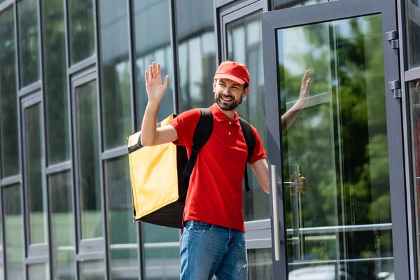 Lächelnder Zusteller mit Thermo-Rucksack winkt in der Nähe eines Gebäudes an der Stadtstraße — Stockfoto