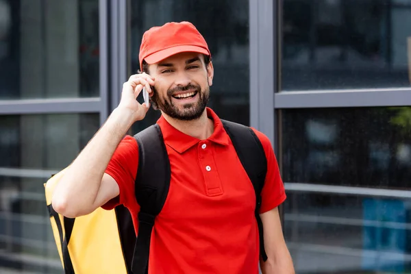 Correio sorridente com mochila térmica falando no smartphone na rua urbana — Fotografia de Stock