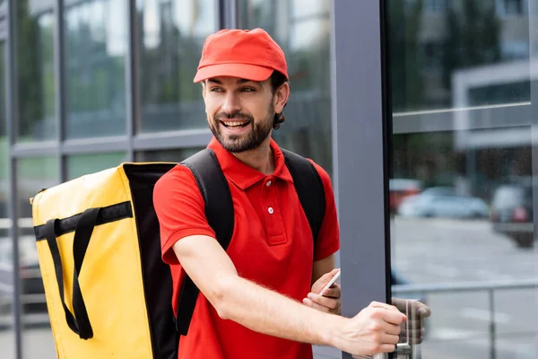 Mensajero sonriente sosteniendo teléfono inteligente cerca de la puerta del edificio en la calle urbana - foto de stock