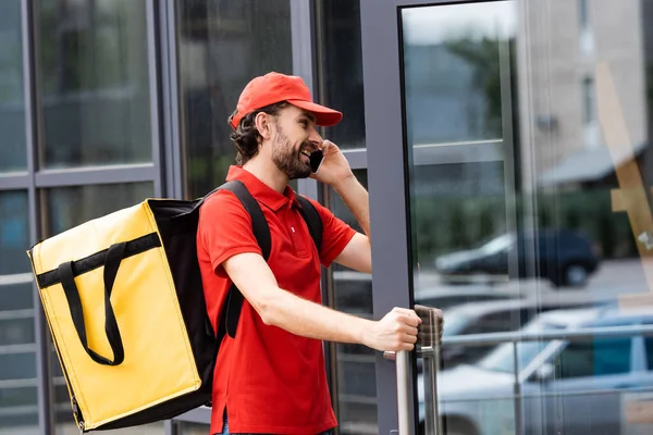 Lächelnder Kurier mit Thermo-Rucksack, der auf dem Smartphone spricht, während er die Tür eines Gebäudes in der städtischen Straße öffnet — Stockfoto