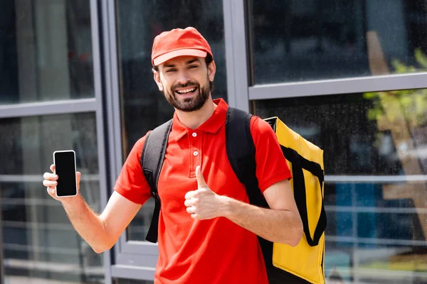 Hermoso mensajero sonriente mostrando como gesto y sosteniendo el teléfono inteligente en la calle urbana - foto de stock