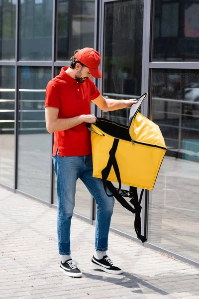 Delivery man in uniform opening thermo bag near building on urban street — Stock Photo
