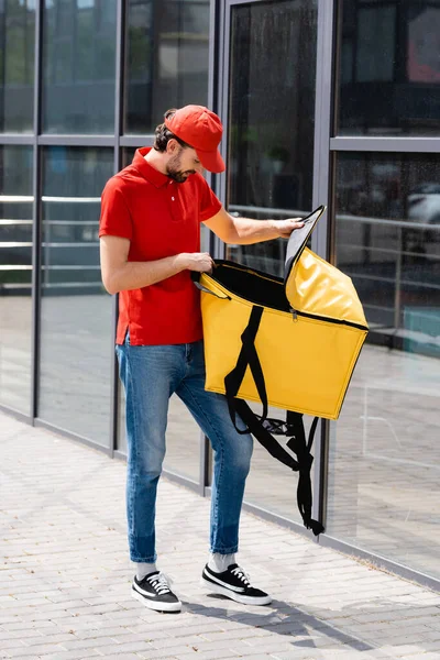 Entrega hombre en uniforme abriendo bolsa de termo cerca del edificio en la calle urbana - foto de stock