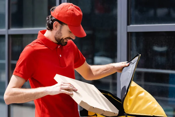 Mensajero sonriente sosteniendo caja de pizza y bolso termo en la calle urbana - foto de stock