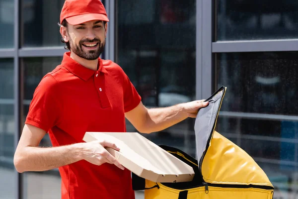 Smiling courier looking at camera while holding pizza box and thermo bag on urban street — Stock Photo