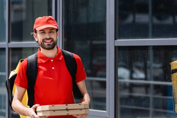 Positive delivery man looking at camera while holding pizza boxes near building on urban street — Stock Photo