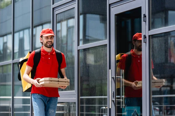 Mensajero sonriente en uniforme sosteniendo cajas de pizza mientras camina cerca del edificio en la calle urbana - foto de stock