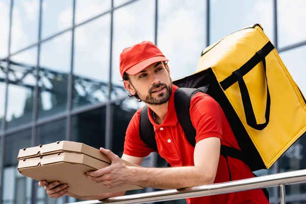 Entrega hombre sosteniendo cajas de pizza cerca de barandilla en la calle urbana - foto de stock