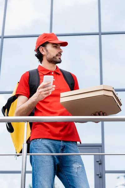 Mensajero guapo sosteniendo cajas de pizza y teléfono inteligente en la calle urbana - foto de stock