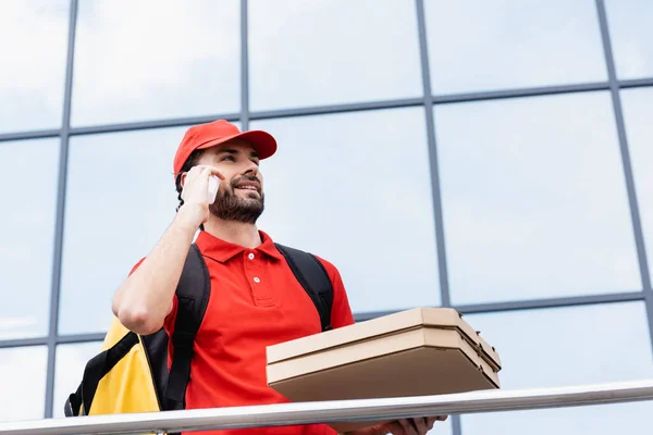 Vista a basso angolo del corriere sorridente che parla sullo smartphone mentre tiene in mano le scatole della pizza sulla strada urbana — Foto stock