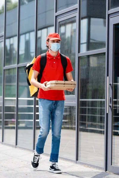 Доставщик в медицинской маске с терморюкзаком с коробками от пиццы во время прогулки по городской улице — стоковое фото
