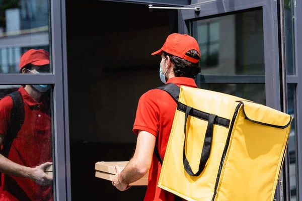 Entrega homem em máscara médica segurando caixas de pizza enquanto de pé perto da porta aberta do edifício — Fotografia de Stock