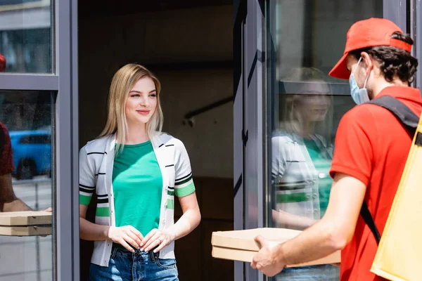 Focus selettivo di donna sorridente in piedi vicino alla porta della costruzione e guardando l'uomo di consegna in maschera medica contenente scatole di pizza — Foto stock