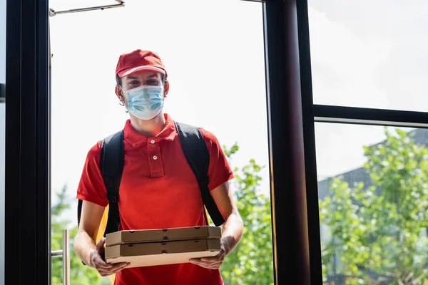 Corriere in maschera medica che tiene scatole di pizza vicino alla porta aperta dell'edificio — Foto stock