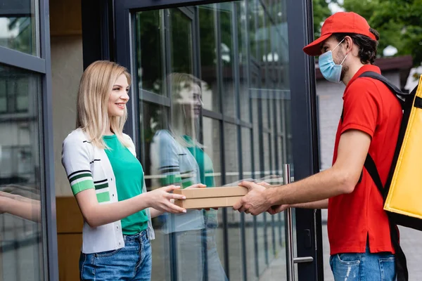 Courier in medical mask giving pizza boxes to smiling woman near building on urban street — Stock Photo