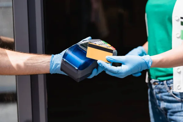 Обрезанный вид женщины в латексных перчатках с кредитной картой рядом с курьером с платежным терминалом на городской улице — стоковое фото