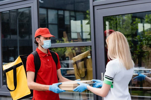 Доставщик в медицинской маске дарит коробки с пиццей женщине в латексных перчатках на городской улице — стоковое фото