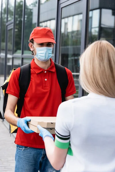 Enfoque selectivo de mensajero en uniforme y máscara médica dando pizza para llevar a la mujer en la calle urbana - foto de stock