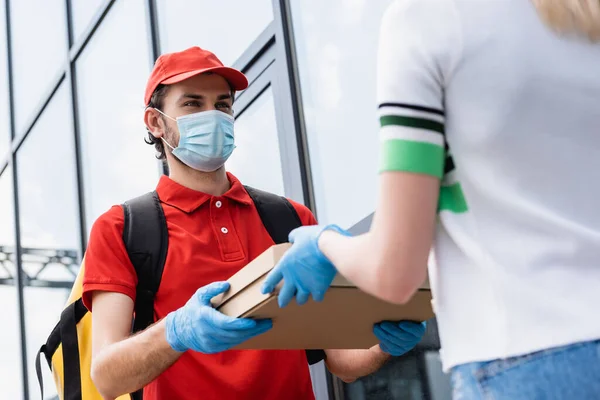 Selective focus of courier in medical mask giving takeaway pizza to girl in latex glove on urban street — Stock Photo