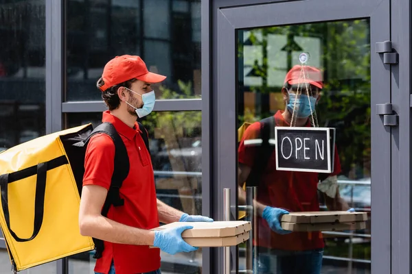 Vista laterale del corriere in maschera medica e guanti in lattice che tengono scatole di pizza vicino al cartello con scritte aperte sulla porta dell'edificio — Foto stock