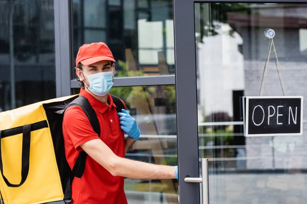 Entrega hombre en máscara médica de pie cerca de la puerta del edificio con letras abiertas en el letrero en la calle urbana - foto de stock