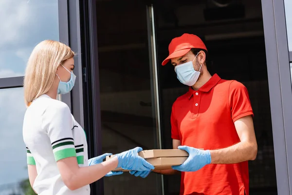 Waiter in medical mask giving pizza boxes to woman in latex gloves near cafe on urban street — Stock Photo