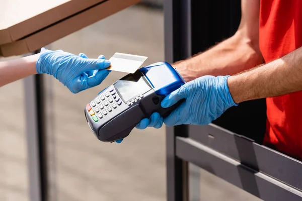 Vista recortada de una mujer sosteniendo cajas de pizza y pagando con tarjeta de crédito cerca del camarero en guantes de látex sosteniendo terminal de pago en la calle urbana - foto de stock