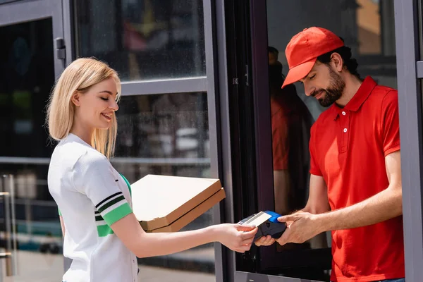 Mujer sonriente sosteniendo cajas de pizza y pagando con paypass cerca del camarero sosteniendo terminal de pago en la calle urbana — Stock Photo