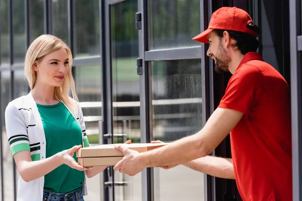 Garçom sorridente em uniforme dando caixas de pizza para jovem mulher perto de café na rua urbana — Fotografia de Stock